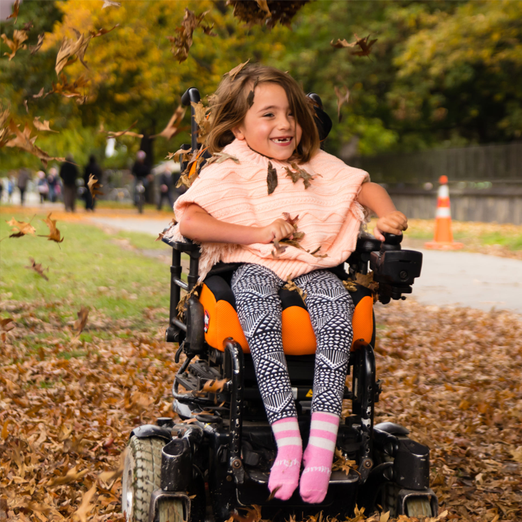 Flicka i rullstol som leker bland höstlöv – rörelseglädje och frihet utomhus.